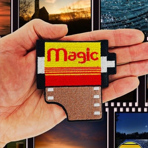 May include: A yellow and red embroidered iron-on patch shaped like a film canister with the word "Magic" in red lettering. The patch has a brown film strip with white perforations.