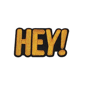 May include: Embroidered patch featuring the word "HEY!" in gold thread on a black backing. The design is simple and bold, with a yellow outline. The exclamation mark is also gold.