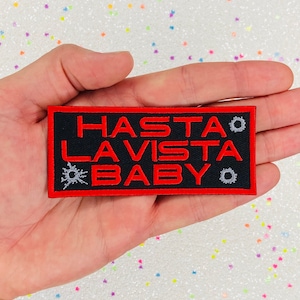 Pode incluir: Um remendo bordado preto e vermelho com o texto "HASTA LA VISTA BABY" em letras vermelhas. O remendo tem uma borda vermelha e dois buracos de bala de cada lado do texto.