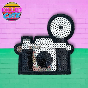 May include: A black and white sequined camera patch with a round lens. The patch has a black background and white sequins. The lens is made of white sequins and has a black center.