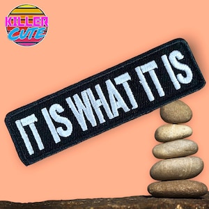 Pode incluir: Remendo de tecido preto com texto branco que diz "IT IS WHAT IT IS". O remendo está encostado em uma pilha de pedras cinzas.