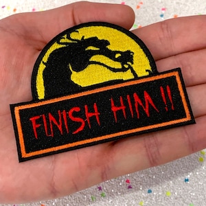 May include: Embroidered patch with a black dragon silhouette on a yellow backdrop, and the red text "FINISH HIM!!". The patch has a black border with an orange outline. Ideal for customising clothing or accessories.