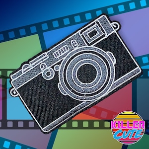 May include: Black and white embroidered iron-on patch of a vintage camera. The camera has a round lens and a rectangular body with a small viewfinder on top.