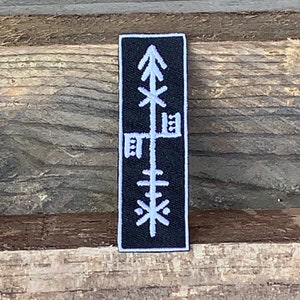 May include: Black embroidered patch featuring a white symbol resembling a stylised tree or plant. The symbol is made up of lines and geometric shapes.