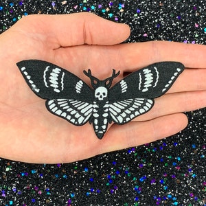 May include: Black and white embroidered iron-on patch of a moth with a skull and antlers. The moth has a detailed wing pattern.