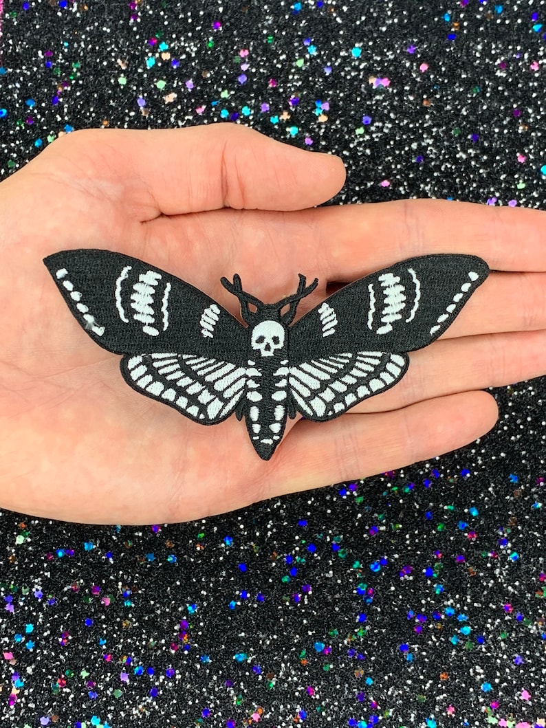 May include: Black and white embroidered iron-on patch of a moth with a skull and antlers. The moth has a detailed wing pattern.