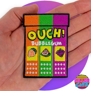 Pode incluir: Um remendo bordado para passar a ferro de uma caixa de pastilha elástica de desenho animado com o texto "OUCH! BUBBLEGUM" e três sabores de fruta diferentes: morango, melancia e uva.