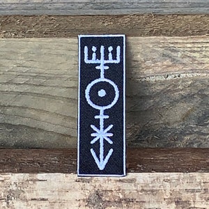 May include: Black embroidered patch with a white symbol. The symbol is a vertical line with a circle at the top, a star in the middle, and an arrow at the bottom.