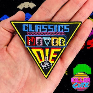 以下が含まれることがあります： 「Classics Never Die」と刺繍されたカラフルなテキストが施された黒い三角形のワッペン。テキストはレトロなアーケードスタイルのフォントです。テキストの下には小さな白いジョイスティックが刺繍されています。