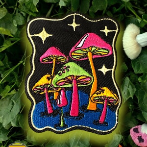 以下が含まれることがあります： 黒地に輝く星を背景に、ピンク、グリーン、イエローのアクセントが施されたカラフルなキノコのグループが刺繍されたパッチ。