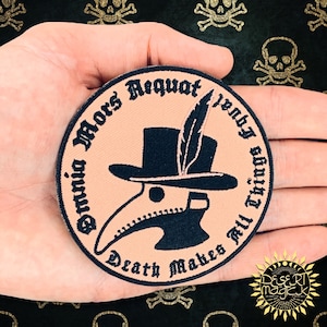 Pode incluir: Um remendo redondo cor de pêssego com bordados pretos. O remendo apresenta um médico da peste estilizado usando um chapéu com uma pena, e o texto "Omnia Mors Aequat" e "Death Makes All Things Equal".