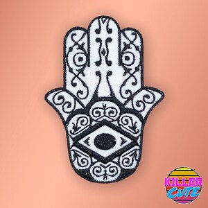 May include: White embroidered hamsa hand with a black eye in the centre. The hamsa is a symbol of protection and good luck.