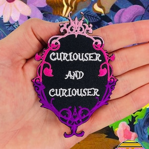 May include: A black and white embroidered patch with a pink and purple frame. The patch features the text "Curiouser and Curiouser" in a white font.