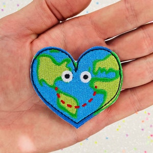 May include: A blue and green embroidered heart-shaped patch with a cartoon-style depiction of the Earth with googly eyes and a red dotted line around the continents.
