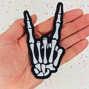 May include: White skeleton hand making the rock and roll hand gesture. The hand is embroidered on a black fabric patch.