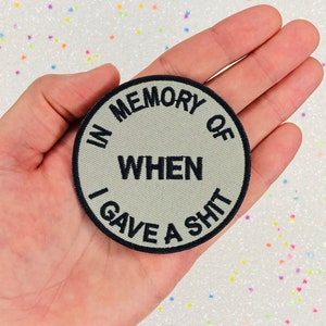 May include: A round, beige patch with black stitching and the text "In memory of when I gave a shit."