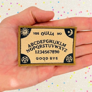 May include: A brown and black embroidered patch with a Ouija board design. The patch features the letters of the alphabet, numbers 1-9 and 0, and the words "YES", "OUIJA", "NO", and "GOOD BYE".
