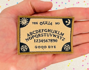 May include: A brown and black embroidered patch with a Ouija board design. The patch features the letters of the alphabet, numbers 1-9 and 0, and the words "YES", "OUIJA", "NO", and "GOOD BYE".
