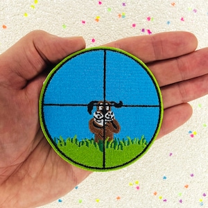 May include: A round, green and blue embroidered patch with a cartoon dog peeking out from behind a green bush. The dog is brown with white eyes and is in the center of a crosshair target.