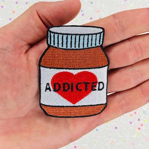 以下が含まれることがあります： 白い蓋と赤いハートに「ADDICTED」と書かれた茶色のガラス瓶。瓶は手に乗っている。