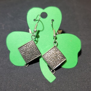May include: A pair of silver-toned, diamond-shaped earrings with intricate embossed patterns. The earrings hang from silver hooks and are displayed on a green shamrock-shaped card. The background is a dark, neutral color.