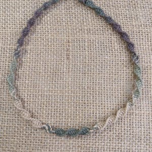 May include: A handmade necklace made of twisted hemp cord in shades of green, brown, and beige. The necklace has a spiral design and is fastened with a wooden bead. The necklace is displayed on a burlap background.