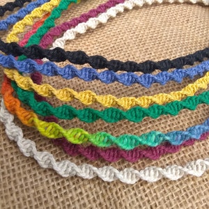 May include: A set of ten colorful crocheted cord necklaces in various colors, including white, beige, green, yellow, blue, and red. The necklaces are arranged in a circle on a brown burlap background.