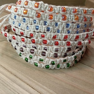 May include: A stack of handmade hemp bracelets with colorful glass beads. The bracelets are made with a macrame knotting technique and feature orange, blue, red, purple, and green beads.