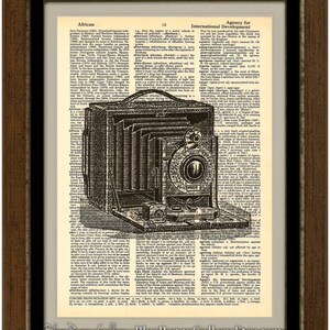 May include: A vintage black and white image of a camera on a page of a dictionary. The camera is a box camera with a bellows and a lens. The dictionary page has text about African countries and the Agency for International Development.