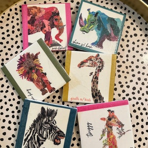 May include: Set of six colorful coasters featuring animal illustrations. The coasters are made of paper and have a textured surface. The animals depicted are a pink elephant, a rhino with a horn, a lion, a giraffe with birds, a zebra, and a giraffe with the word "twiggy" written on the coaster.