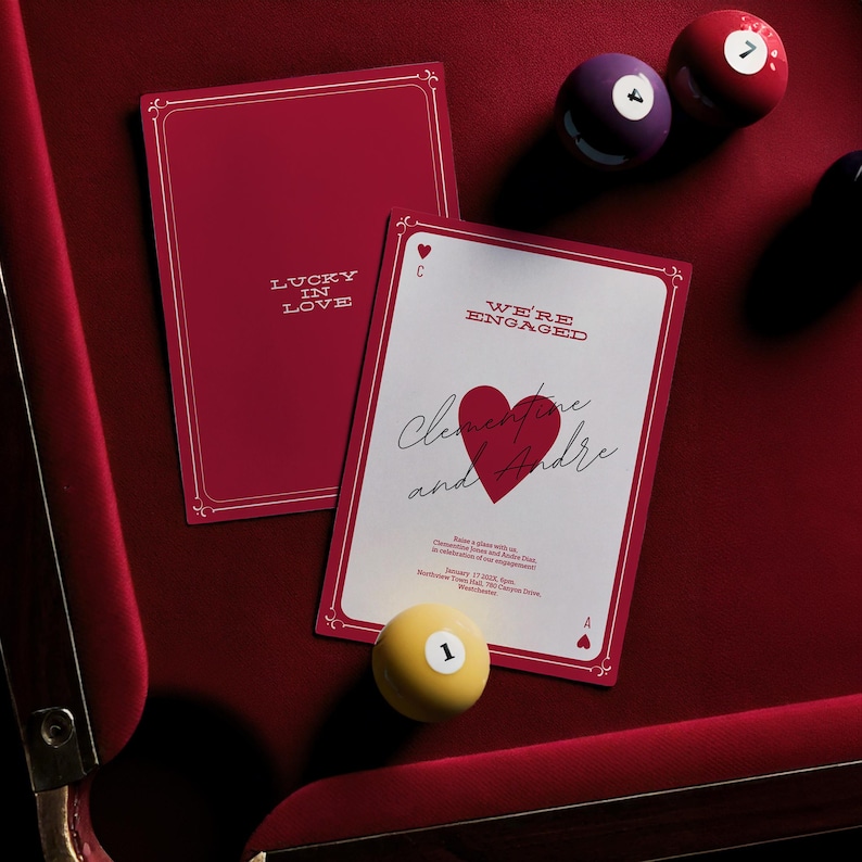 K&ouml;nnte beinhalten: Eine rote und wei&szlig;e Verlobungsank&uuml;ndigungskarte mit einem roten Herzen und dem Text "We're Engaged" und "Clementine and Andre". Die Karte liegt auf einem roten Filz mit Billardkugeln.