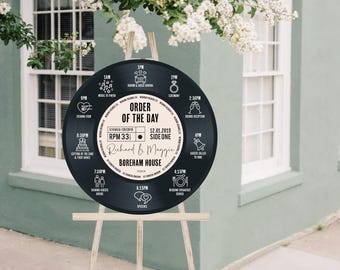 May include: A black vinyl record wedding sign on a wooden easel. The sign displays the wedding schedule, including times and events, with the names Richard & Maggie and the location Boreham House. White flowers are in the background.