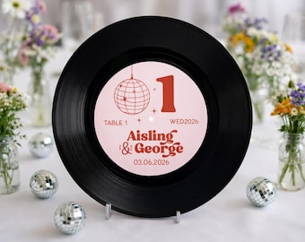 May include: A black vinyl record with a pink and white center label. The label features a disco ball illustration, the number 1, and the names "Aisling & George" with the wedding date 03.06.2026. Decorative disco balls and flowers are in the background.