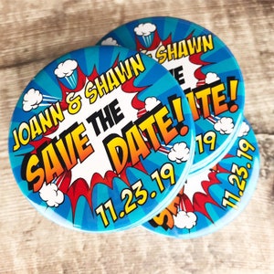 May include: A stack of round blue and red comic book style save the date buttons with the text "JOANN & SHAWN SAVE THE DATE! 11.23.19" in yellow and white lettering.