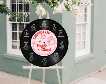 May include: A black vinyl record wedding sign on a wooden easel. The sign displays the wedding schedule with the text "Order of the Day" and the names "Paige & Chad" along with the date, March 26, 2027. The sign is decorated with pink accents.