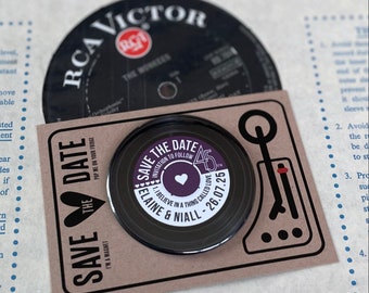 May include: Save the date magnet with a black and white record design. The text reads "Save the Date" with a heart and "Invitation to Follow" in a circle. The date is "26.07.25" and the names "Elaine & Niall" are printed below. The text "I Believe in a Thing Called Love" is printed in a smaller font.