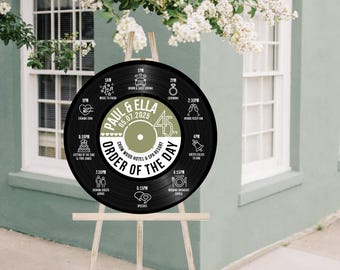 May include: A black vinyl record-shaped wedding sign on a wooden easel, displaying the order of events for Paul & Ella's wedding on 05.07.2025. The sign includes event times and icons, set against a backdrop of a building and white flowers.