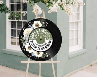 May include: A black vinyl record with a white floral design and the text "SARA & LEWIS" and "JUNE 21, 2025" and "WELCOME TO OUR WEDDING" and "ALL YOU NEED IS LOVE" on it. The record is on a wooden easel.