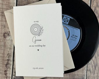 May include: A white folded greeting card with black text that reads to my Groom on our wedding day and the date 19.06.2020. The card features a line art illustration of a vinyl record with leaves and a small heart icon, placed over a vinyl record.