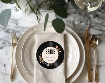May include: A white napkin with a black vinyl record place card on top. The record has a floral design and the text "BRIDE" with the names "Richard & Maggie" and the date "21.08.2023".