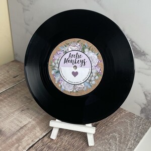 May include: A black vinyl record with a brown and white label featuring a floral design and the text "Arctic Monkeys" in a purple heart. The label also includes the text "Wedding Records Ltd."