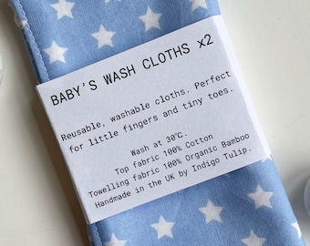 May include: Two blue baby washcloths with white stars. The washcloths are made of 100% cotton and 100% organic bamboo. They are machine washable at 30 degrees Celsius. "BABY'S WASH CLOTHS x2 Reusable, washable cloths. Perfect for little fingers and tiny toes. Wash at 30°C. Top fabric 100% Cotton Towelling fabric 100% Organic Bamboo Handmade in the UK by Indigo Tulip."