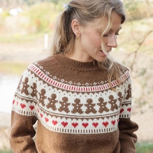 May include: A brown knitted sweater with a round neckline. The sweater features a pattern of gingerbread men and red hearts on a white background. The top of the sweater has a red and white striped design.