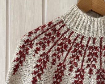 Puede incluir: Un suéter de punto de color crema con un diseño estampado en rojo y crema. El suéter tiene un cuello alto y está hecho de un material suave y cálido.