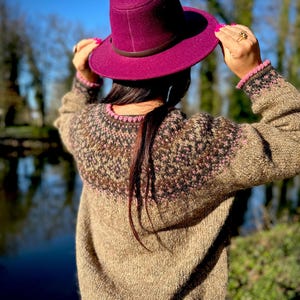 May include: A magenta felt fedora hat with a dark brown band paired with a long-sleeved, beige knit sweater featuring a circular yoke pattern in shades of brown, black, and pink, with matching pink trim at the neckline and sleeve cuffs.