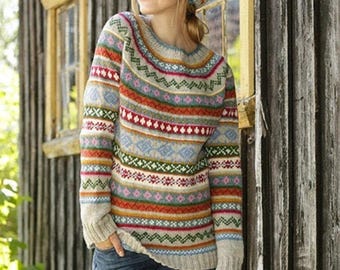 Puede incluir: Una mujer lleva un suéter de punto colorido con un patrón geométrico y un gorro de punto a juego. El suéter tiene rayas de rojo, naranja, amarillo, verde, azul y blanco. El gorro es de estilo beanie.