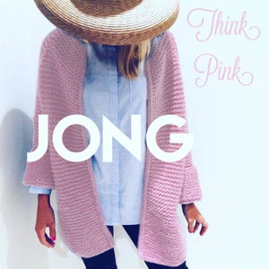 Puede incluir: Un cárdigan de punto rosa con un frente amplio y abierto y mangas largas. El cárdigan se lleva sobre una camisa de botones azul claro y pantalones negros. La modelo lleva un sombrero de paja y el texto "Think Pink" y "JONG" es visible en la imagen.