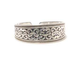 May include: A silver cuff bracelet with an intricate, ornate design. The bracelet is made of a smooth, polished metal and has a slightly curved shape.