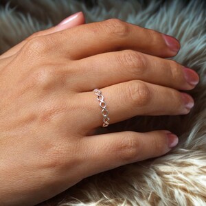 Puede incluir: Un anillo plateado con un diseño de símbolo de infinito. El anillo está hecho de metal fino y presenta una serie de bucles interconectados. El anillo se lleva en un dedo. El fondo es una superficie suave y texturizada.