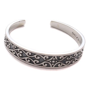Könnte beinhalten: Ein silbernes Armreif-Armband mit offenem Design. Das Armband hat ein kompliziertes, sich windendes Muster entlang des Bandes. Das Metall hat eine polierte Oberfläche, und das Design ist mit dunklen Akzenten versehen. Es ist ein Schmuckstück.